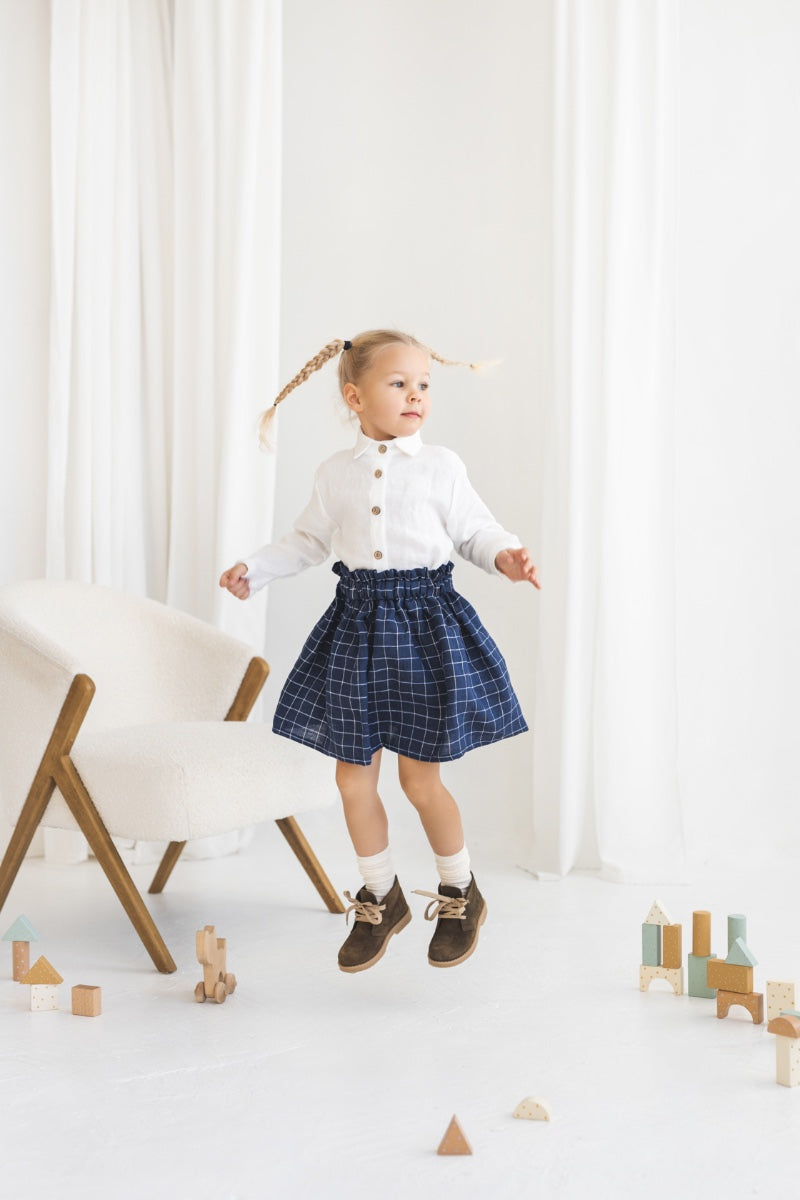 Young girl in a white blouse and blue plaid linen skirt standing in a minimalistic room.