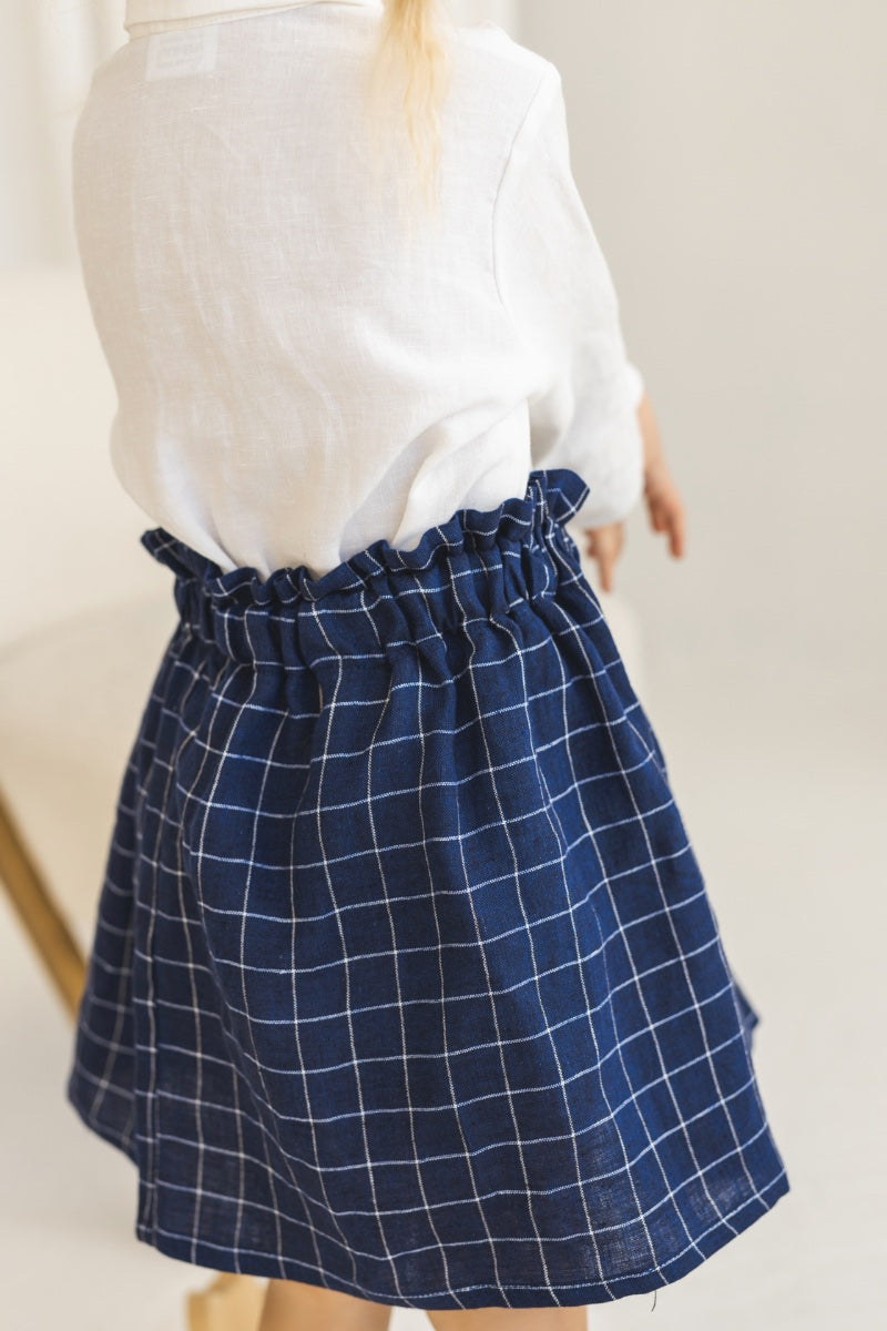 Young girl in a white blouse and blue plaid linen skirt standing in a minimalistic room.