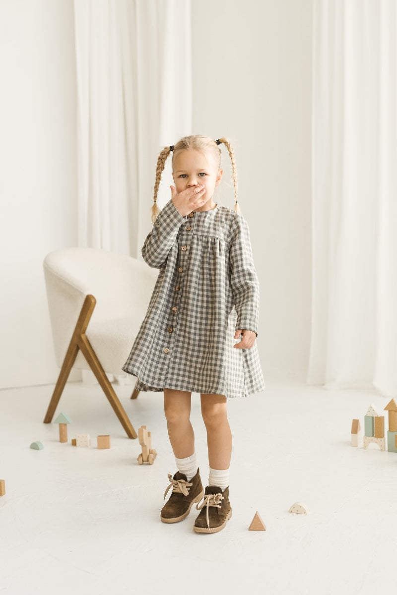 Young girl in a grey linen checkered dress standing in a minimalistic room with white curtains and wooden toys.