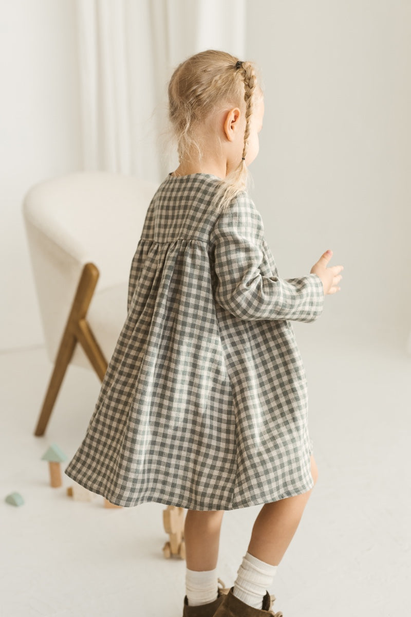 Young girl in a grey linen checkered dress standing in a minimalistic room with white curtains and wooden toys.