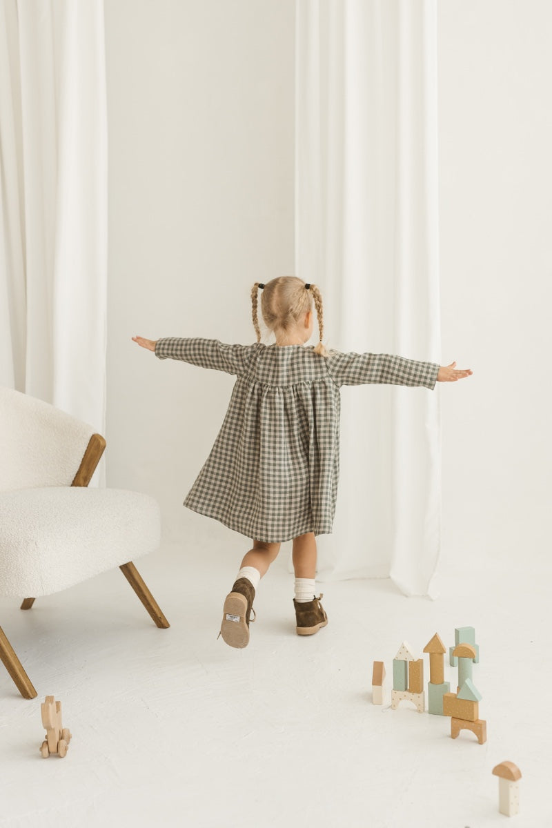 Young girl in a grey linen checkered dress standing in a minimalistic room with white curtains and wooden toys.