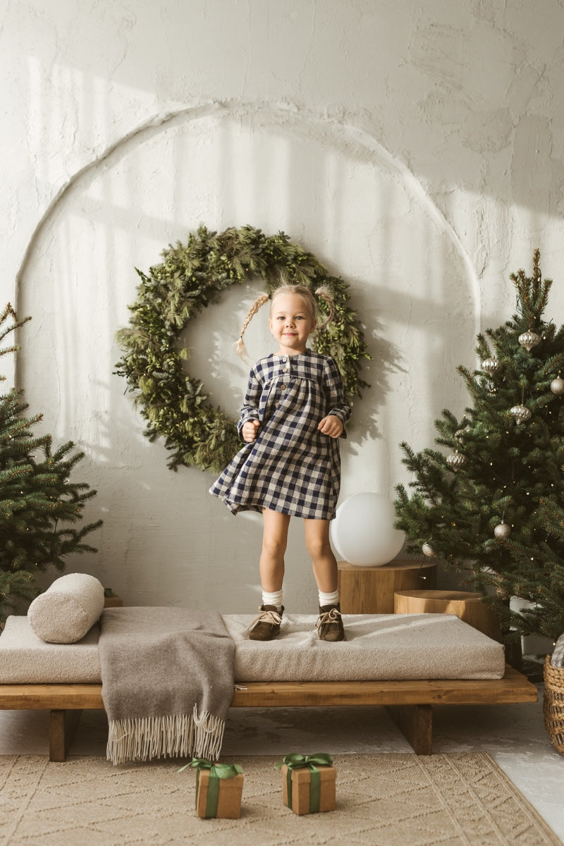 Young girl in a checkered linen dress standing in a minimalistic room with white curtains and wooden toys on the floor.