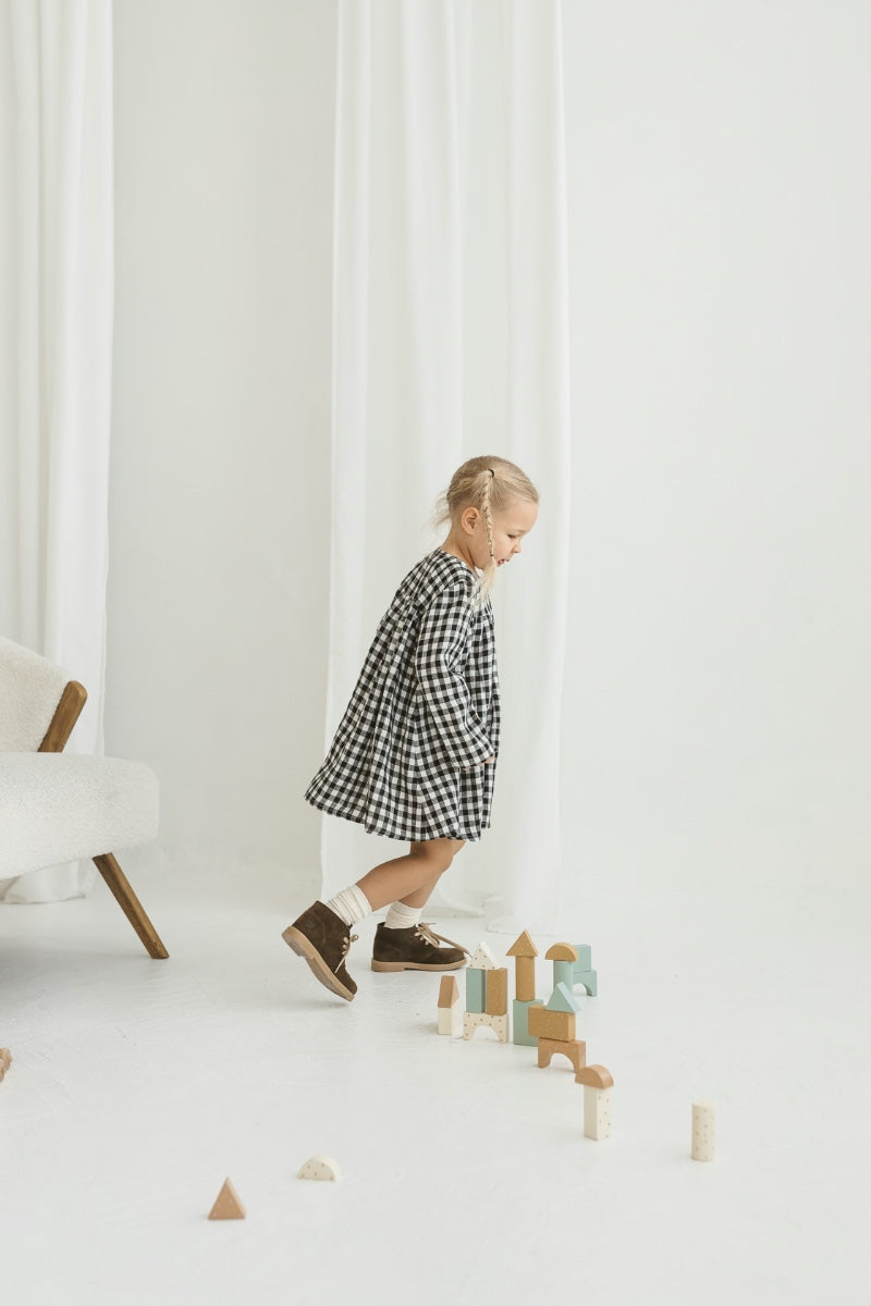 Child in a checkered linen dress standing on a white floor with wooden toys.
