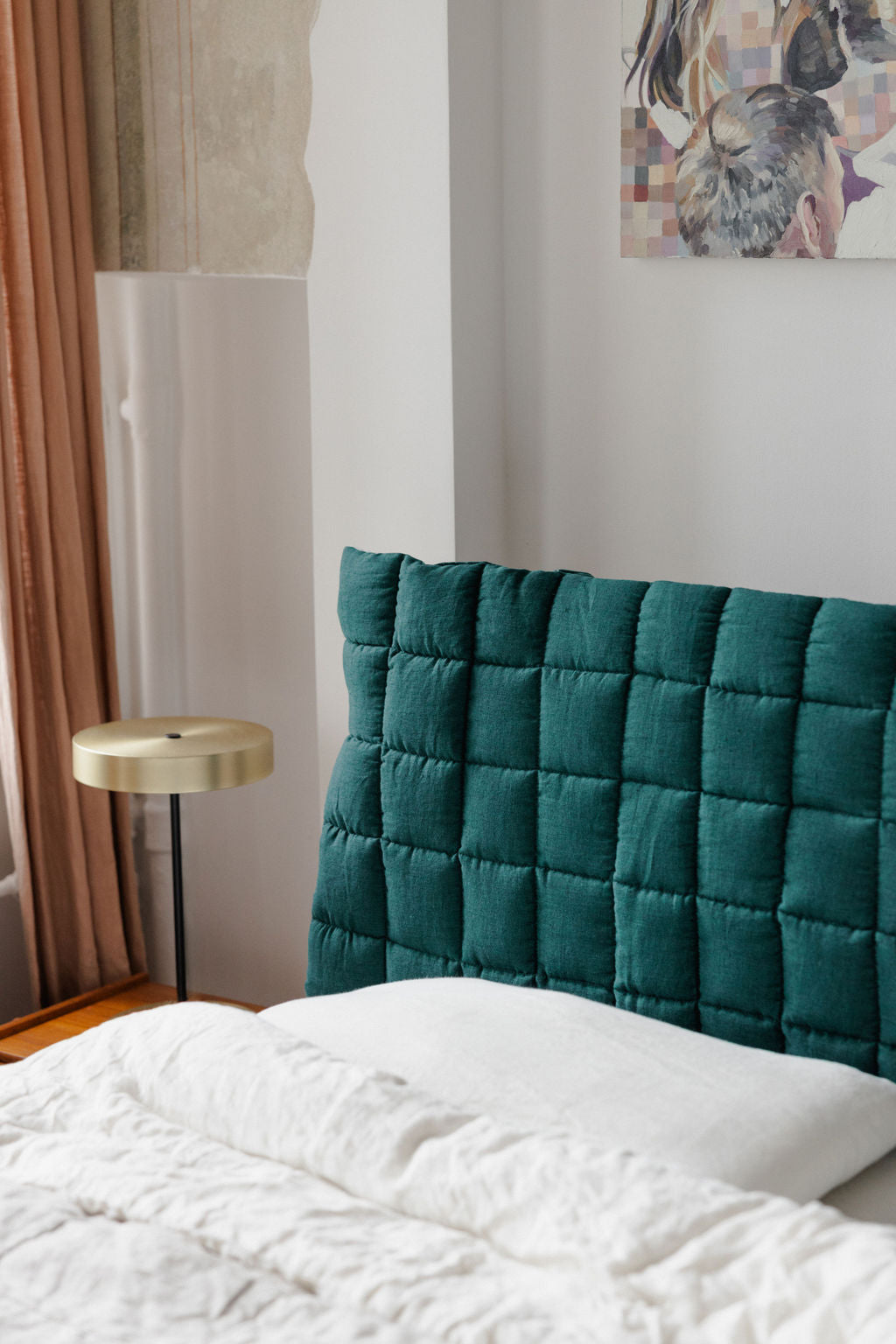 Teal quilted headboard against a white wall with a small round wooden table and abstract art in the background.
