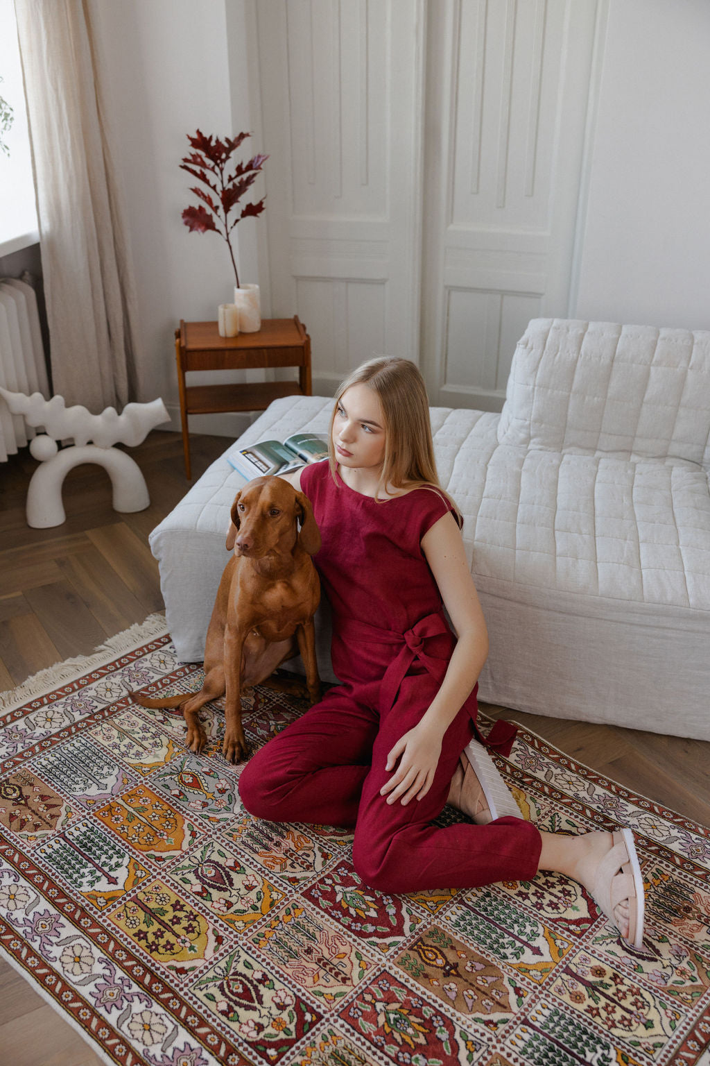 Woman in a red outfit sitting on a patterned rug with a dog in a living room.