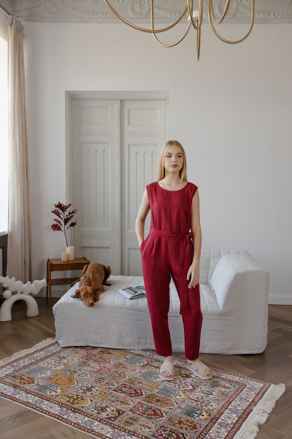 Woman in a red jumpsuit standing in a room with a dog on a bed and a chandelier.