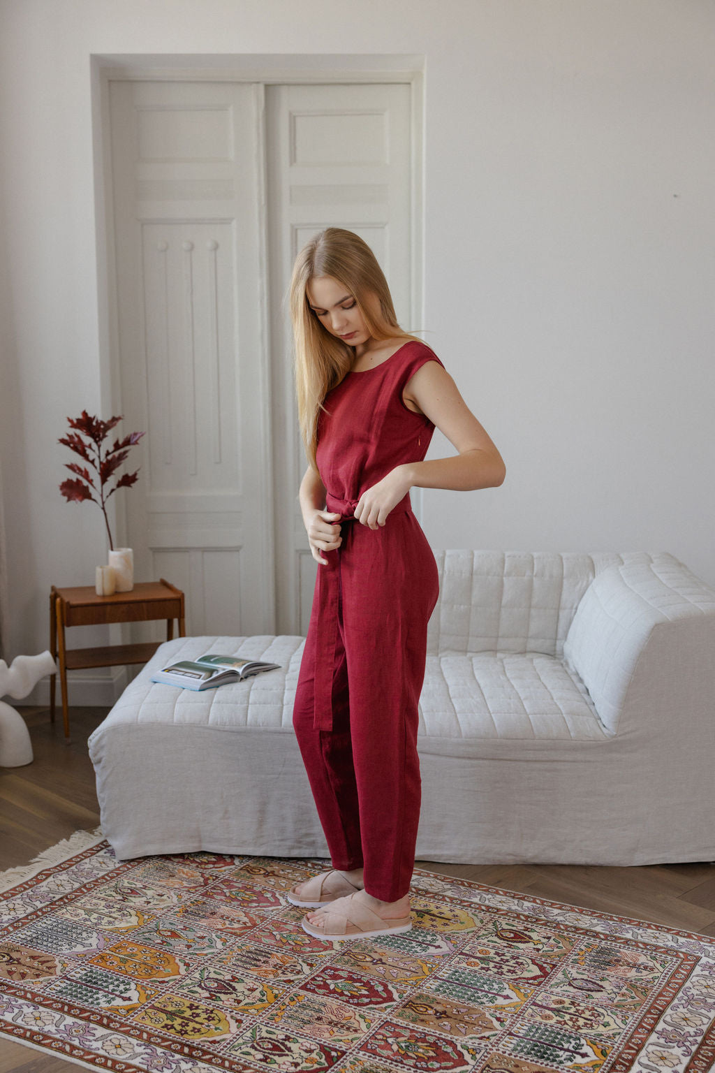 Woman in a red jumpsuit standing in a room with a white sofa and decorative rug.