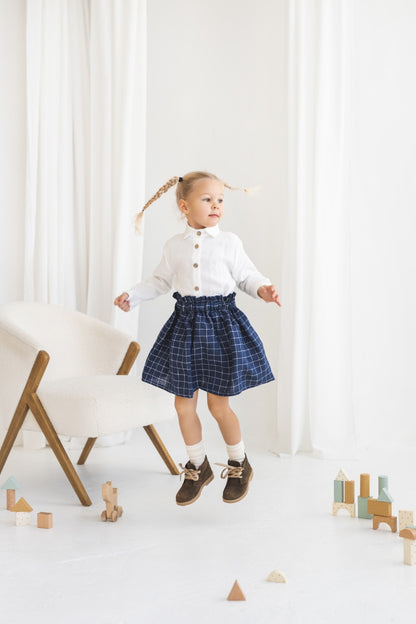Young girl in a white blouse and blue plaid linen skirt standing in a minimalistic room.
