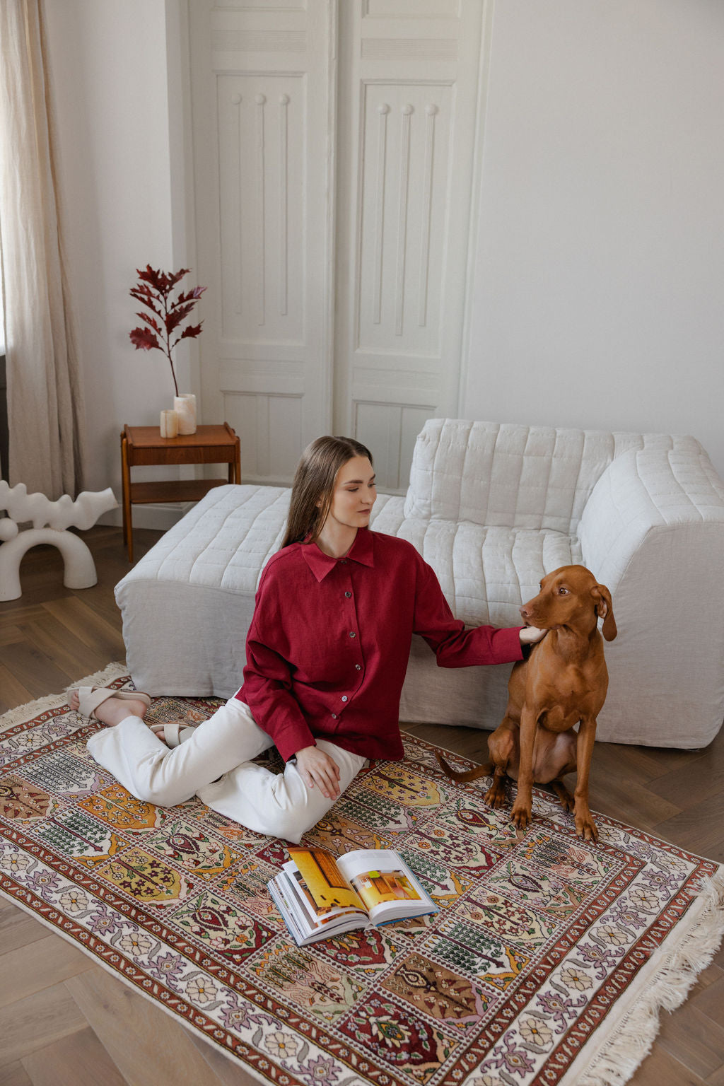 Woman in red shirt and white pants sitting on a patterned rug with a dog, in a room with a white sofa and wooden side table.