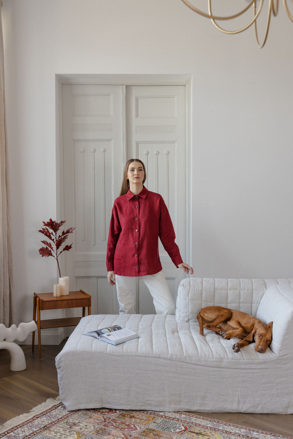 Woman in a red shirt standing in a living room with a dog on a couch.