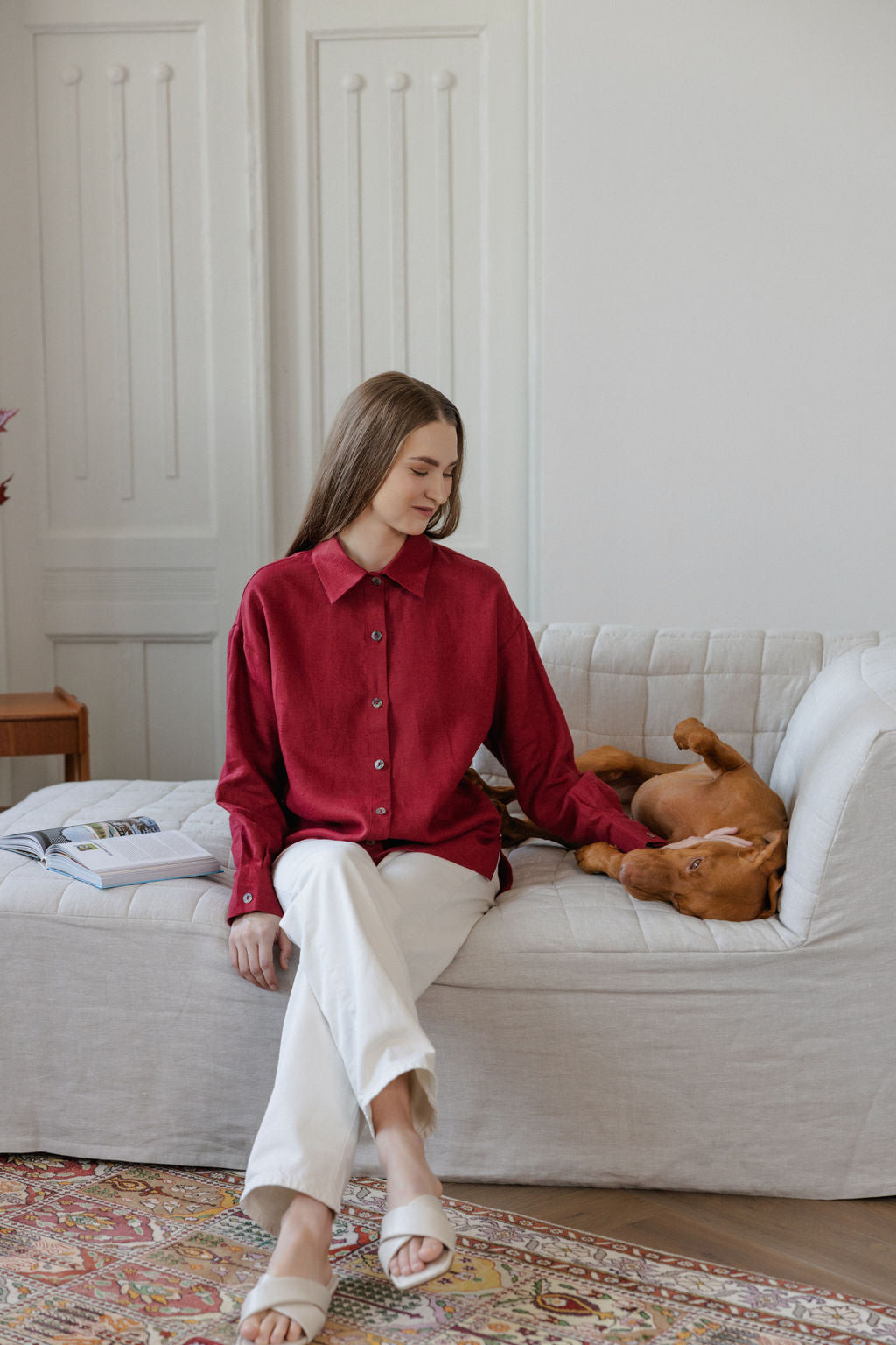 Woman in a red shirt sitting on a white couch with a dog, in a room with a patterned rug and wooden side table.