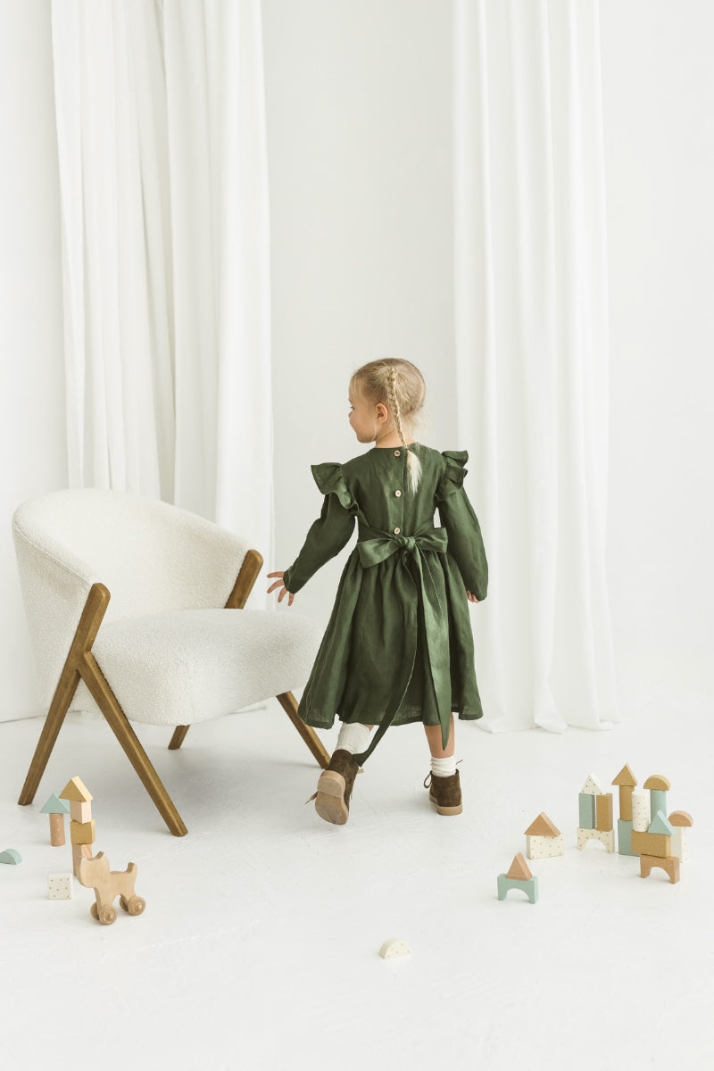 Child in a green  linendress standing near a white chair with wooden legs on a white floor.

