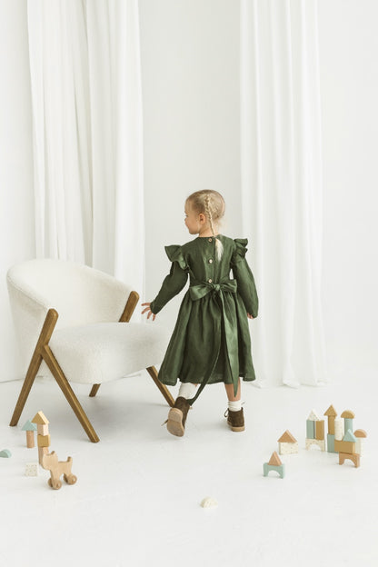 Child in a green  linendress standing near a white chair with wooden legs on a white floor.

