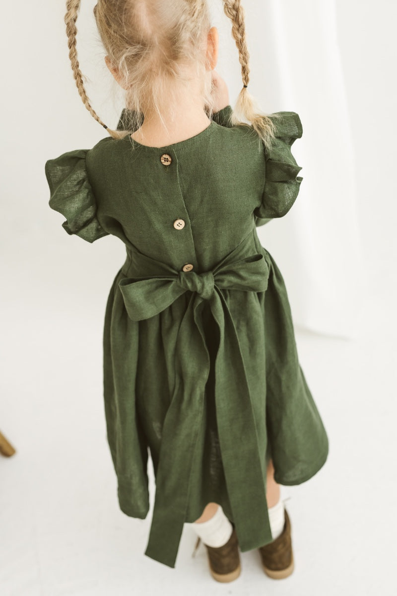 Child in a green dress standing near a white chair with wooden legs on a white floor.

