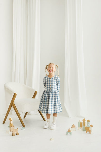 Girl in a blue checkered linen dress standing in a room with white curtains and wooden toys on the floor.

