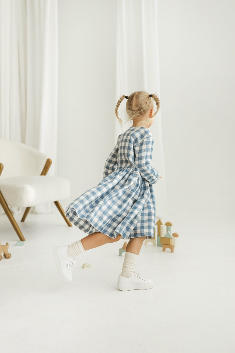 Girl in a blue checkered linen dress standing in a room with white curtains and wooden toys on the floor.
