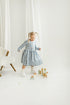 Child in a blue checkered linen dress standing in a minimalistic room with white curtains and wooden toys.

