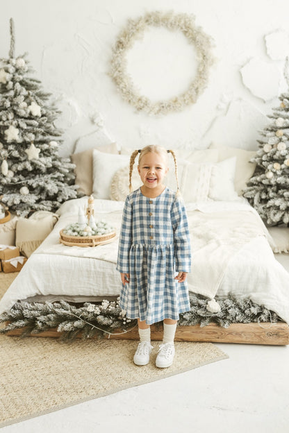 Girl in a blue checkered linen dress standing in a decorated room with Christmas trees and wreaths.

