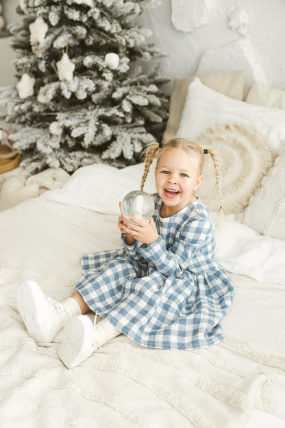 Child in a blue checkered linen dress holding a silver ornament in front of a decorated Christmas tree.


