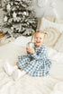 Child in a blue checkered linen dress holding a silver ornament in front of a decorated Christmas tree.

