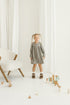 Young girl in a grey linen checkered dress standing in a minimalistic room with white curtains and wooden toys.
