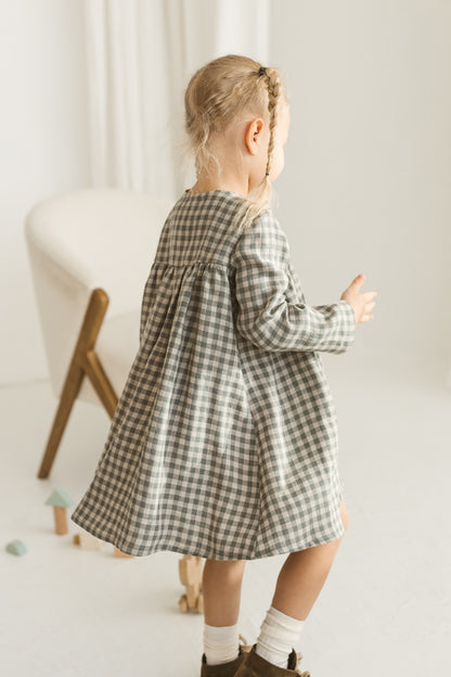 Young girl in a grey linen checkered dress standing in a minimalistic room with white curtains and wooden toys.
