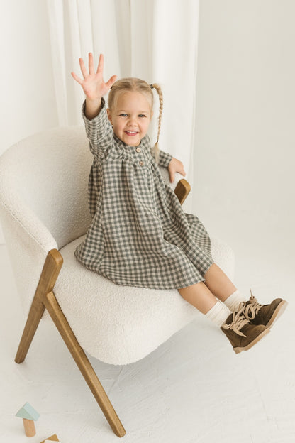 Young girl in a grey linen checkered dress standing in a minimalistic room with white curtains and wooden toys.
