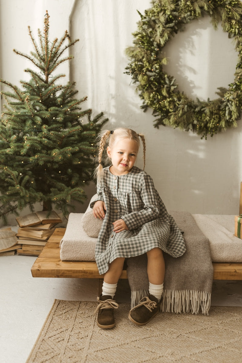 Young girl in a grey linen checkered dress standing in a minimalistic room with white curtains and wooden toys.
