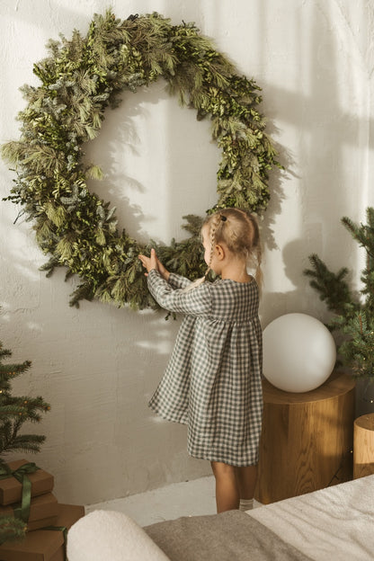 Young girl in a grey linen checkered dress standing in a minimalistic room with white curtains and wooden toys.
