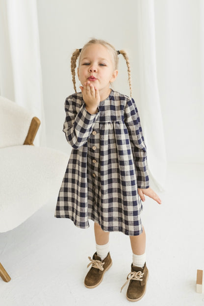 Young girl in a checkered linen dress standing in a minimalistic room with white curtains and wooden toys on the floor.
