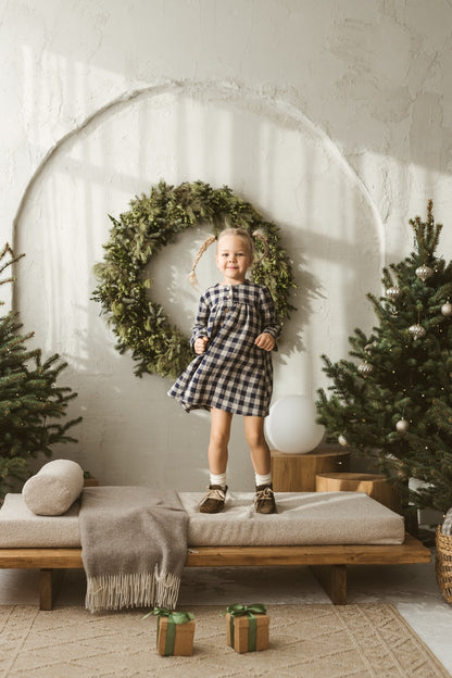 Young girl in a checkered linen dress standing in a minimalistic room with white curtains and wooden toys on the floor.
