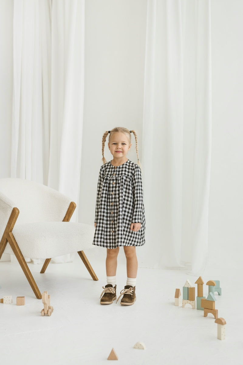 Child in a checkered linen dress standing on a white floor with wooden toys.
