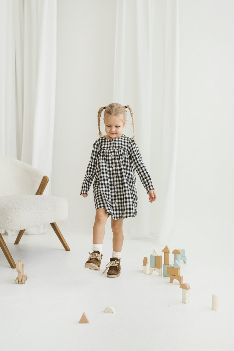 Child in a checkered linen dress standing on a white floor with wooden toys.


