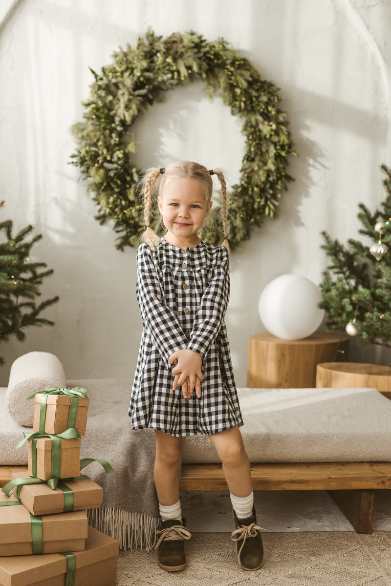 Child in a checkered linen dress standing on a white floor with wooden toys.
