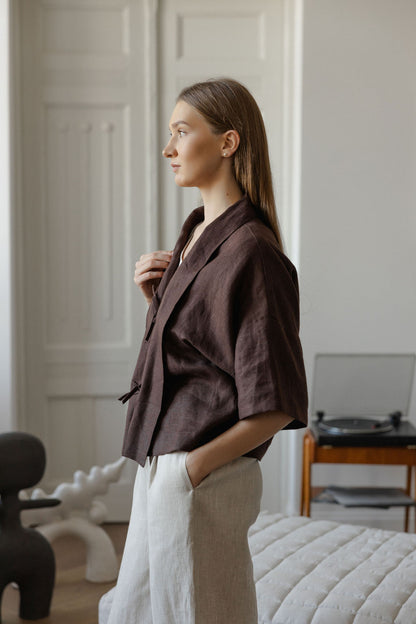 Woman wearing a brown  linen blazer and white pants in a room with a bed and chair.