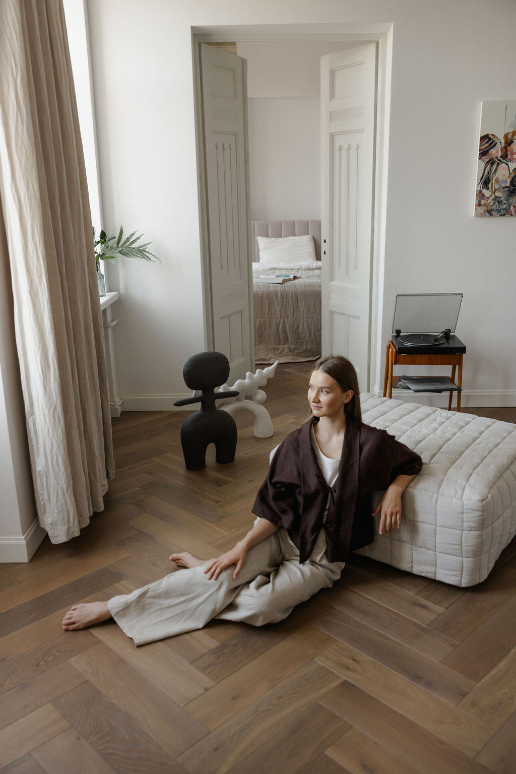 Woman sitting on a white ottoman in a room with wooden flooring and a mirror.