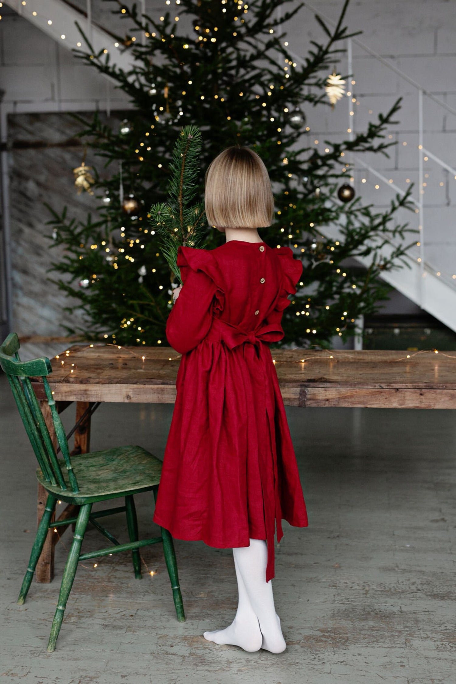 Young girl in a linen red dress holding a Christmas tree branch in front of a decorated Christmas tree.