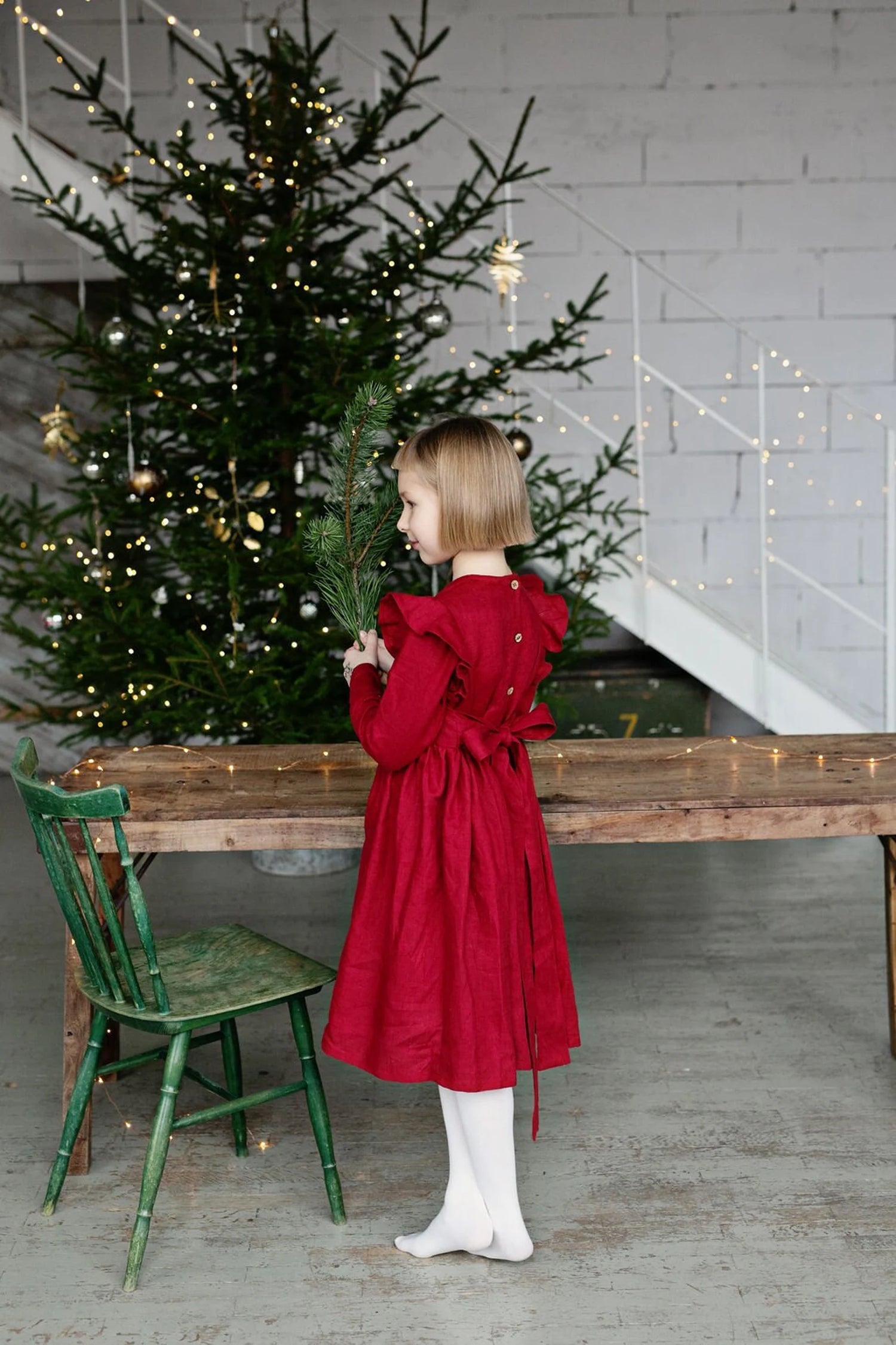 Young girl in a red linen dress standing near a decorated Christmas tree indoors.