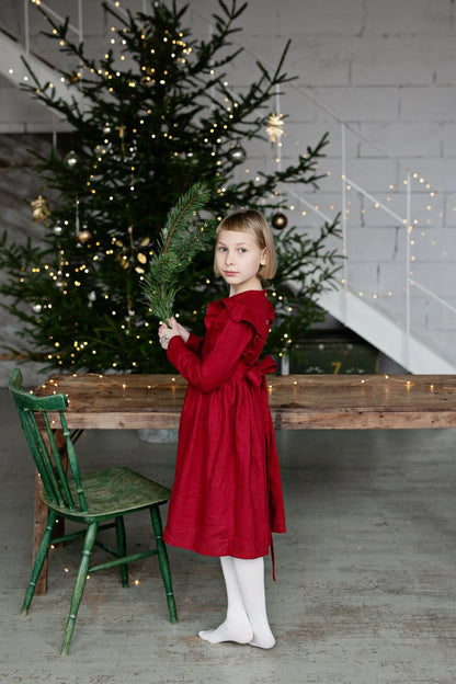 Young girl in a linen red dress holding a Christmas tree branch in front of a decorated Christmas tree.