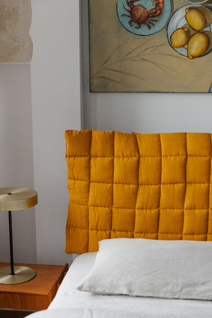 Yellow quilted headboard on a bed with a side table and artwork in the background.