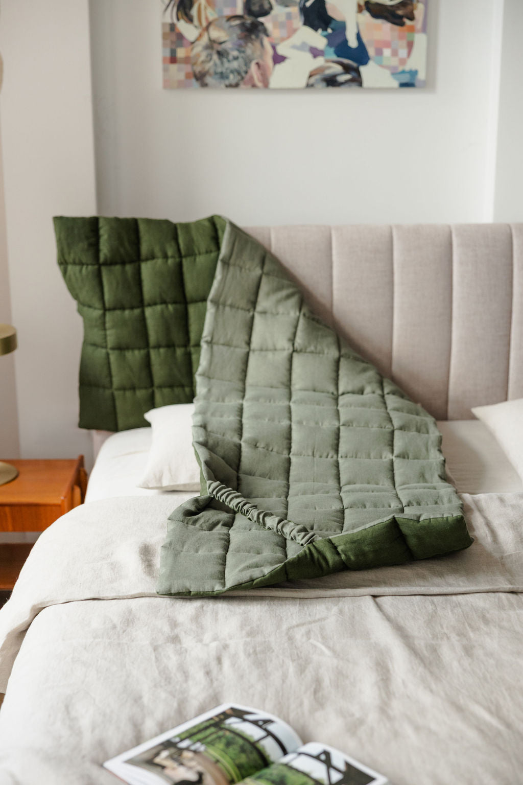 Green weighted blanket draped over a bed with a colorful abstract painting on the wall.