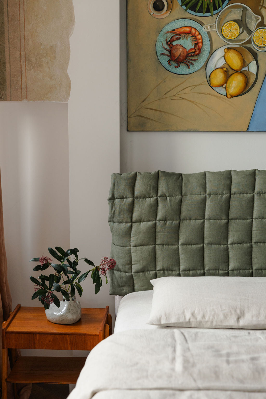 Bedroom with a green quilted headboard, white bedding, and a wooden nightstand with a plant.