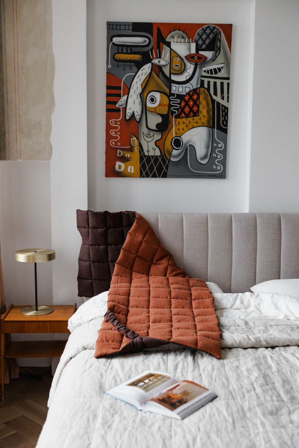 Bedroom with a bed featuring a quilt and pillows, a nightstand with a lamp, and a colorful abstract painting on the wall.