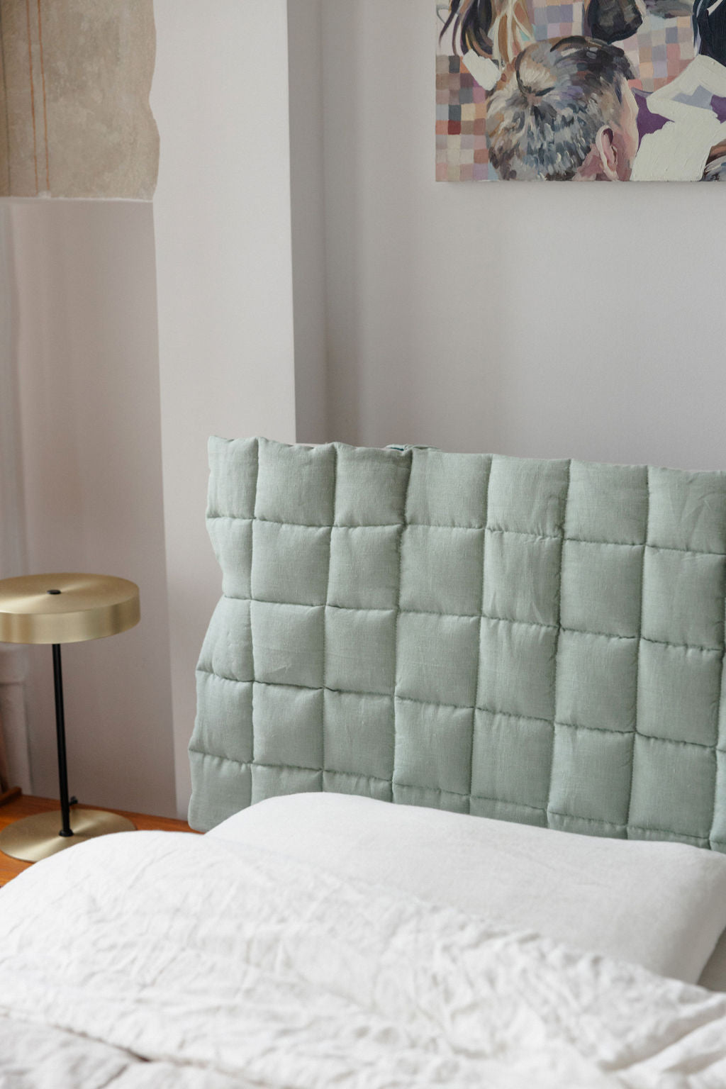 Light green quilted headboard against a white wall with a small table and abstract art in the background.