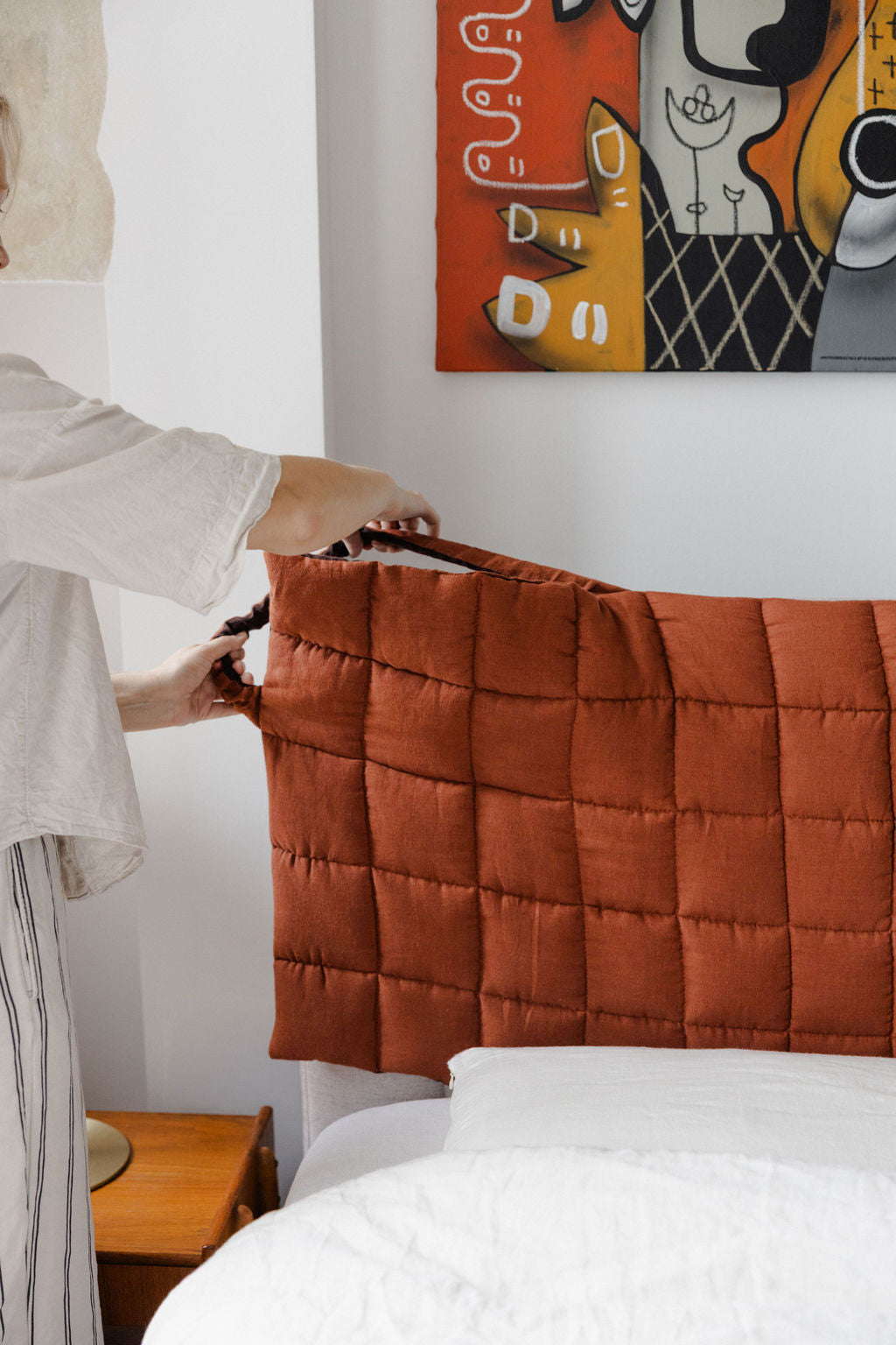 Person adjusting a brown quilted headboard on a bed with a colorful abstract painting in the background.