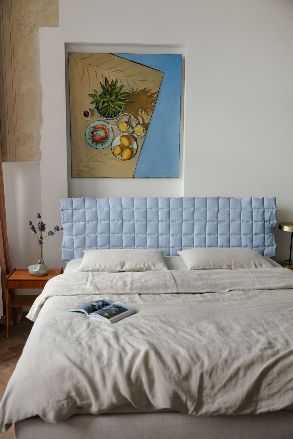 Bedroom with a bed featuring a light blue headboard and beige bedding, with a painting on the wall.
