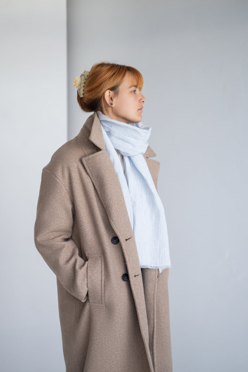Person wearing a beige coat and light blue linen scarf against a plain background

