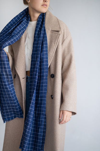 Person wearing a beige coat and blue checkered linen scarf against a white background